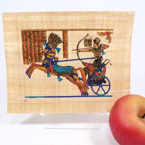 May include: An Egyptian papyrus art print depicting a chariot scene with a horse, archer, and hieroglyphs. The artwork features vibrant colors, including blue, orange, and gold, on a beige background. The print is displayed on a clear stand.