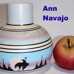 May include: A hand-painted ceramic vase with a blue, white, and pink design. The vase features a silhouette of a Native American riding a horse in front of a full moon. The vase is signed "Ann Navajo".