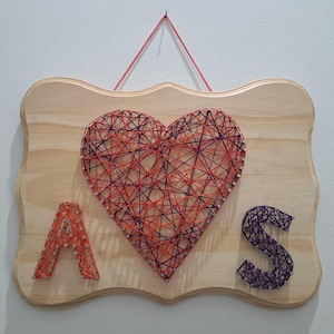 Peut inclure: Une plaque en bois avec un motif en fil tendu représentant un cœur en fils orange et violet, encadré par les lettres A et S. La lettre A est formée de fil orange, tandis que la lettre S est en fil violet. Une ficelle rose permet de suspendre l'objet.