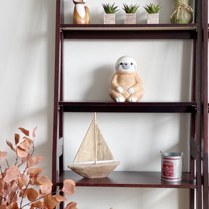 Puede incluir: Una estanter&iacute;a de madera de color marr&oacute;n oscuro con un letrero colgante que dice LITTLE MAN CAVE, una figura de zorro, tres suculentas peque&ntilde;as, una figura de perezoso, un velero de madera, un cubo de metal y cajas tejidas.