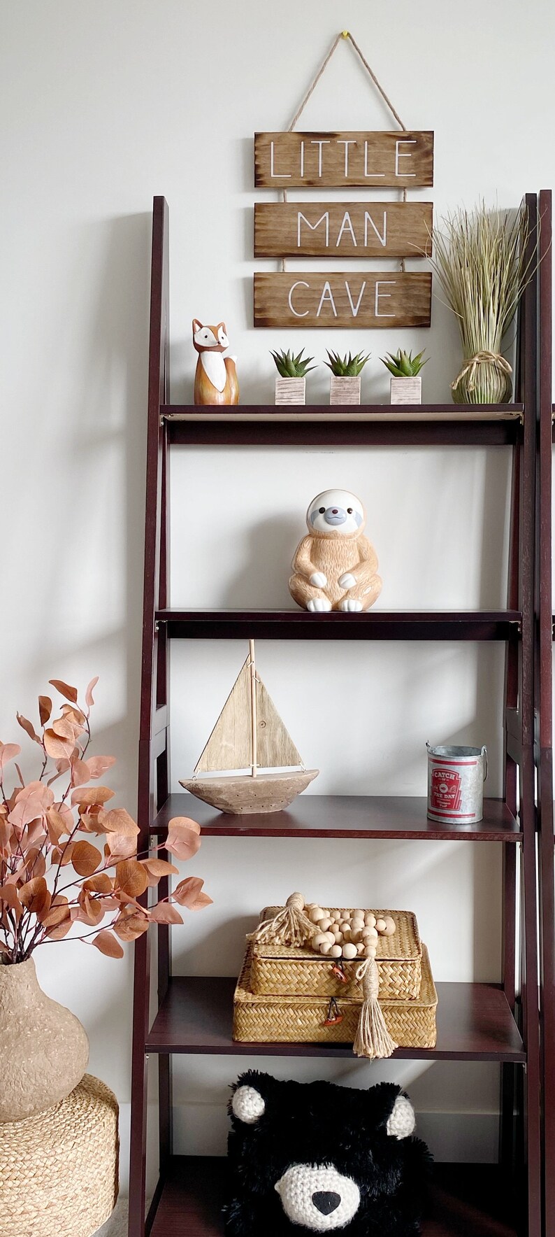 Puede incluir: Una estanter&iacute;a de madera de color marr&oacute;n oscuro con un letrero colgante que dice LITTLE MAN CAVE, una figura de zorro, tres suculentas peque&ntilde;as, una figura de perezoso, un velero de madera, un cubo de metal y cajas tejidas.