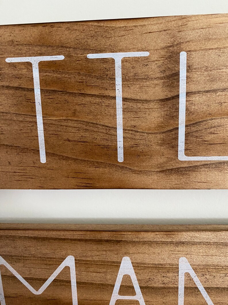 Puede incluir: Una vista de cerca de dos tablones de madera apilados con un acabado de grano de madera marr&oacute;n natural. El tabl&oacute;n superior presenta las letras blancas T, T y L, mientras que el tabl&oacute;n inferior muestra las letras blancas M, A y N en fuente.
