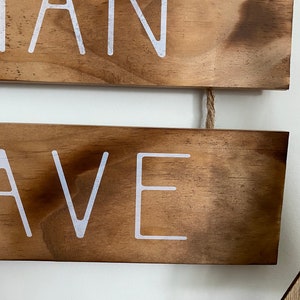 Puede incluir: Dos tablones de madera horizontales con un acabado te&ntilde;ido de color marr&oacute;n oscuro, conectados por un trozo de cuerda de yute. El tabl&oacute;n superior presenta el texto blanco "IAN" y el inferior el texto blanco "AVE" en una fuente sans-serif.