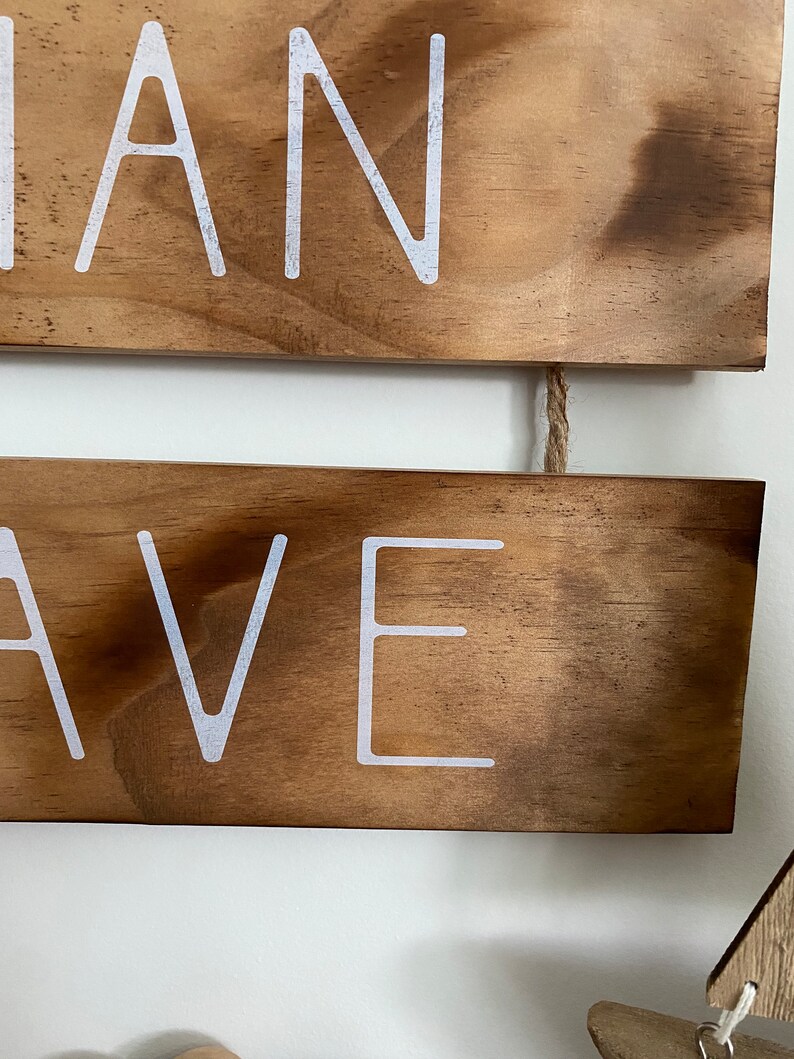 Puede incluir: Dos tablones de madera horizontales con un acabado te&ntilde;ido de color marr&oacute;n oscuro, conectados por un trozo de cuerda de yute. El tabl&oacute;n superior presenta el texto blanco "IAN" y el inferior el texto blanco "AVE" en una fuente sans-serif.
