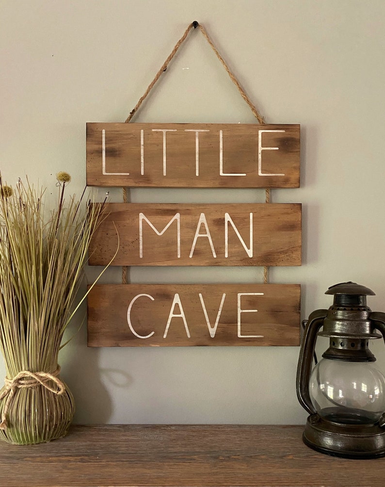 Puede incluir: Un letrero de pared colgante compuesto por tres tablones de madera marr&oacute;n horizontales unidos por cordel. El texto "LITTLE MAN CAVE" est&aacute; pintado en letras may&uacute;sculas blancas sobre los tablones. El letrero tiene un acabado desgastado.