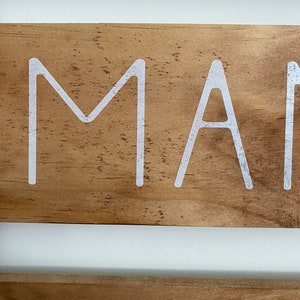 Puede incluir: Un letrero de madera con veta de color marr&oacute;n claro y letras blancas. La secci&oacute;n superior muestra las letras M y A en una fuente sans-serif limpia. La secci&oacute;n inferior muestra las letras G, A y V en la misma tipograf&iacute;a blanca sans-serif.