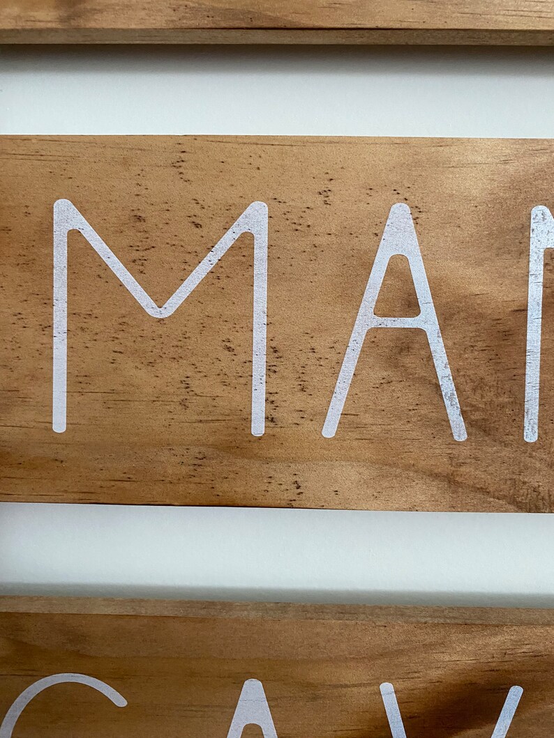 Puede incluir: Un letrero de madera con veta de color marr&oacute;n claro y letras blancas. La secci&oacute;n superior muestra las letras M y A en una fuente sans-serif limpia. La secci&oacute;n inferior muestra las letras G, A y V en la misma tipograf&iacute;a blanca sans-serif.