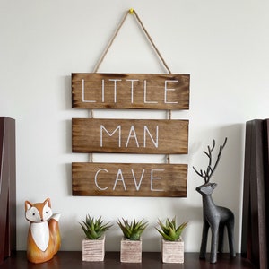 Puede incluir: Un letrero de madera colgante compuesto por tres tablones horizontales te&ntilde;idos en un acabado marr&oacute;n oscuro. El texto "LITTLE MAN CAVE" est&aacute; pintado en letras may&uacute;sculas blancas sobre los tablones. El letrero cuelga de una cuerda de yute.