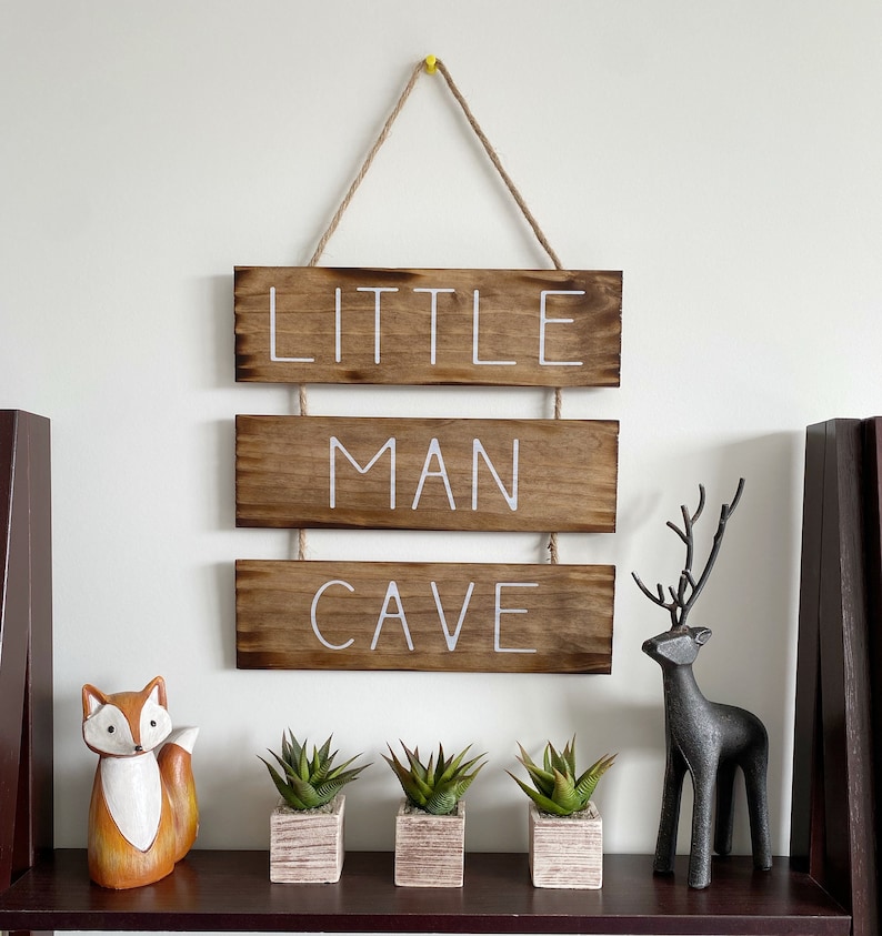 Puede incluir: Un letrero de madera colgante compuesto por tres tablones horizontales te&ntilde;idos en un acabado marr&oacute;n oscuro. El texto "LITTLE MAN CAVE" est&aacute; pintado en letras may&uacute;sculas blancas sobre los tablones. El letrero cuelga de una cuerda de yute.