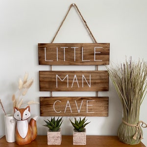 Puede incluir: Un letrero de pared colgante compuesto por tres tablones de madera horizontales te&ntilde;idos de marr&oacute;n oscuro conectados por cordel. El texto LITTLE MAN CAVE est&aacute; impreso en letras may&uacute;sculas blancas sobre las tablas. Acabado de veta de madera.