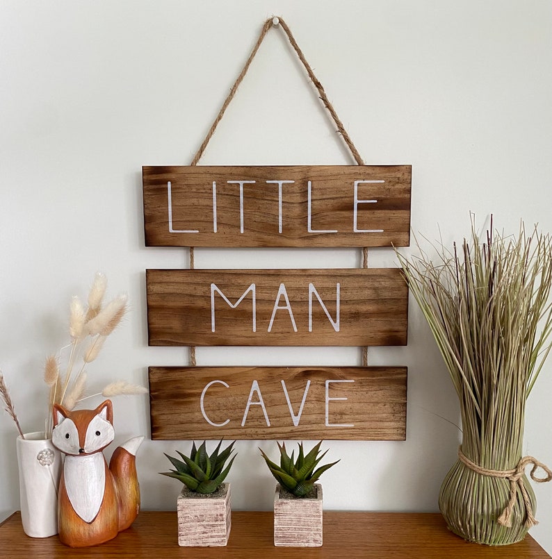 Puede incluir: Un letrero de pared colgante compuesto por tres tablones de madera horizontales te&ntilde;idos de marr&oacute;n oscuro conectados por cordel. El texto LITTLE MAN CAVE est&aacute; impreso en letras may&uacute;sculas blancas sobre las tablas. Acabado de veta de madera.