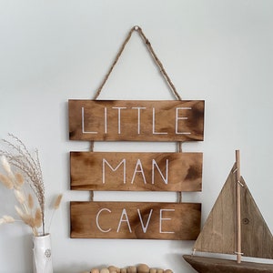 Puede incluir: Un letrero de pared colgante compuesto por tres tablones de madera horizontales te&ntilde;idos de oscuro unidos por cordel. El texto blanco en los tablones dice LITTLE MAN CAVE. El letrero se muestra con un modelo de velero de madera y un jarr&oacute;n.