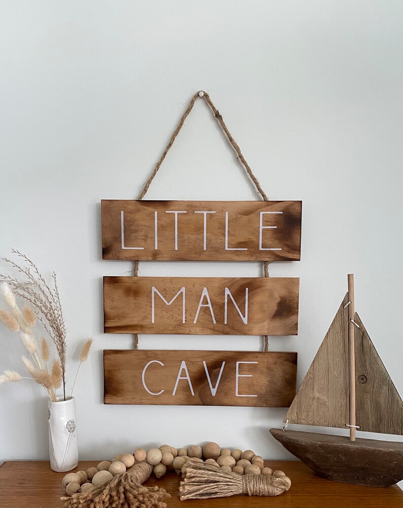 Puede incluir: Un letrero de pared colgante compuesto por tres tablones de madera horizontales te&ntilde;idos de oscuro unidos por cordel. El texto blanco en los tablones dice LITTLE MAN CAVE. El letrero se muestra con un modelo de velero de madera y un jarr&oacute;n.