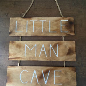 Puede incluir: Un letrero de madera colgante que consta de tres tablones rectangulares conectados por cordel. La madera tiene un acabado marr&oacute;n te&ntilde;ido con bordes m&aacute;s oscuros. El texto blanco dice LITTLE en el tabl&oacute;n superior, MAN en el medio y CAVE abajo.