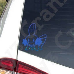May include: A blue vinyl decal of a Mudkip character applied to the rear window of a white vehicle. The sticker features the distinct outline of the creature with its large head fin, side gills, and small feet, contrasting against the dark glass.