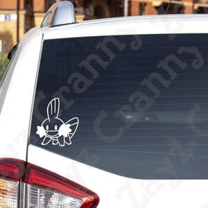 May include: A white vinyl decal of the Mudkip character from the Pok&eacute;mon franchise applied to the rear window of a white vehicle. The graphic features the creature's distinct head fin, wide cheeks, and small body in a simple line art style.