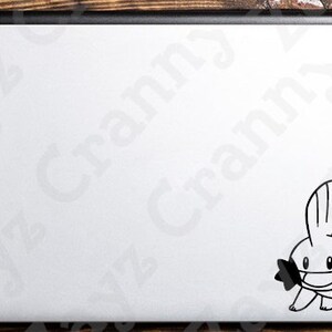 May include: A white laptop lid featuring a black vinyl decal of the character Mudkip. The character is depicted in a simple line art style with a prominent fin on its head, large eyes, and distinct cheek structures, positioned in the bottom right corner.
