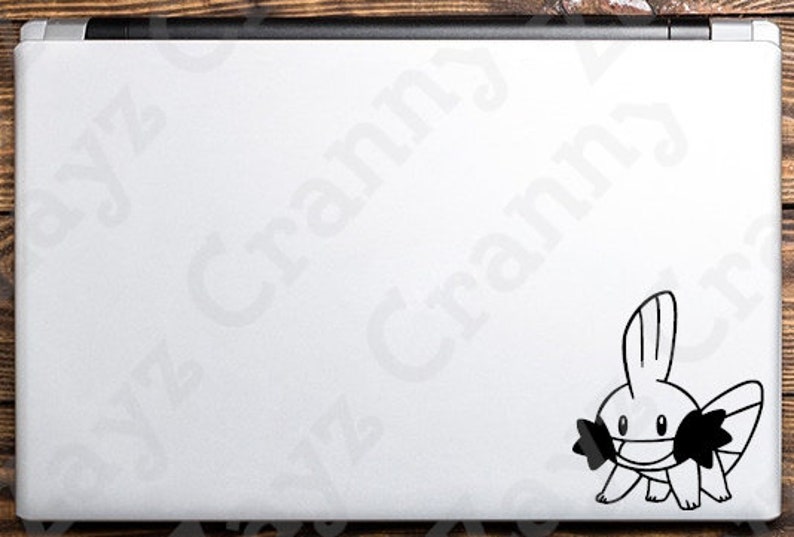 May include: A white laptop lid featuring a black vinyl decal of the character Mudkip. The character is depicted in a simple line art style with a prominent fin on its head, large eyes, and distinct cheek structures, positioned in the bottom right corner.
