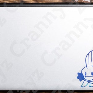 May include: A silver laptop lid featuring a blue vinyl decal of a Mudkip character. The decal is positioned in the bottom right corner of the laptop surface. The laptop has a black hinge at the top edge and a smooth, metallic silver finish.