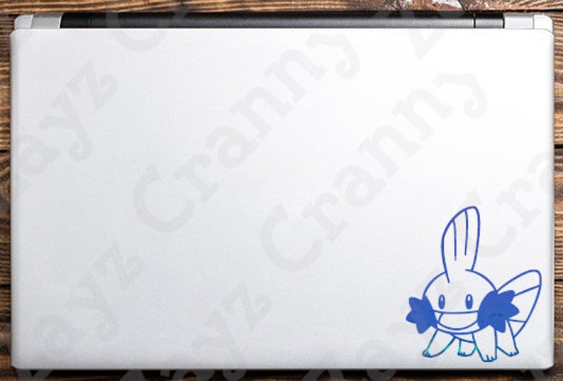 May include: A silver laptop lid featuring a blue vinyl decal of a Mudkip character. The decal is positioned in the bottom right corner of the laptop surface. The laptop has a black hinge at the top edge and a smooth, metallic silver finish.