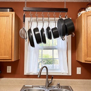 May include: A dark wood ceiling-mounted pot rack suspended by silver chains, featuring eight metal hooks holding various black non-stick pans and a silver mesh strainer. The rack is positioned above a stainless steel kitchen sink and faucet.