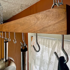 May include: A wooden pot rack suspended from the ceiling by chains. The rack has several metal hooks for hanging pots and pans. The wood is a warm brown color, and the hooks are silver. A window with a lace curtain is visible in the background.