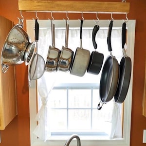 May include: A kitchen pot rack with various stainless steel pots, pans, and a colander hanging from hooks. The rack is made of wood and suspended from the ceiling by chains. The background includes a window and orange wall.