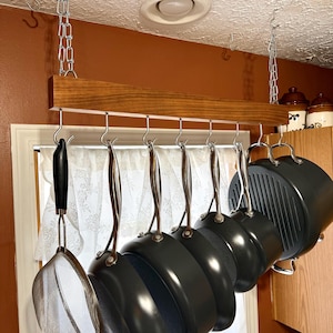 May include: A rectangular wooden pot rack suspended from the ceiling by two silver metal chains. The rack features eight silver metal hooks holding a variety of black cookware, including a mesh strainer and several round pans with long handles.