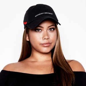 Puede incluir: Una gorra de béisbol negra con el texto "HEARTBREAKER X MONEYMAKER" bordado en la parte delantera. La gorra tiene un parche de corazón rojo en el lateral.