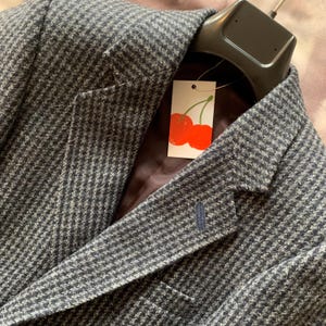 Könnte beinhalten: Ein dunkelgrauer und blauer Hahnentritt-Blazer mit fallendem Revers und einem Zwei-Knopf-Verschluss. Der Blazer hat ein kleines blaues Knopfdetaill am Revers und ein Etikett mit Kirschdesign.