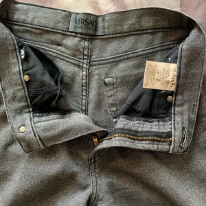 May include: Close-up of gray Versace Jeans Couture denim jeans. The jeans are unbuttoned and unzipped, revealing the interior pockets and a gold zipper. The Versace label is visible at the waistband.