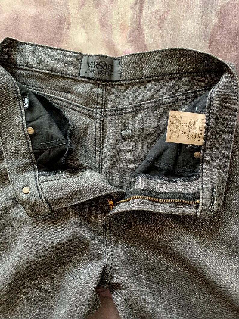 May include: Close-up of gray Versace Jeans Couture denim jeans. The jeans are unbuttoned and unzipped, revealing the interior pockets and a gold zipper. The Versace label is visible at the waistband.