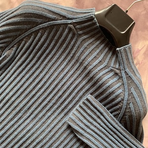 May include: Close-up of a dark gray ribbed knit sweater with blue stripes. The sweater has a high neckline and long sleeves. The fabric is a soft, textured knit, and the design is a classic, timeless style.