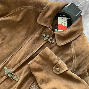 May include: A brown suede jacket with a classic design. The jacket features a collar, toggle closures, and a tag with a cherry design. The jacket is a warm, neutral color, and the details are visible.