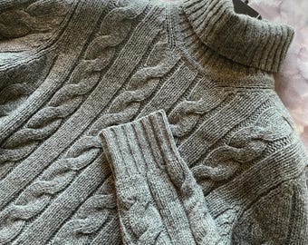 May include: A gray cable-knit turtleneck sweater. The sweater features a textured cable-knit design on the body and sleeves, with a ribbed turtleneck collar and cuffs. The sweater is made of a soft, warm material, perfect for cooler weather.