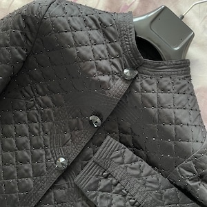 Könnte beinhalten: Schwarze Steppjacke mit Stehkragen und dekorativen Knöpfen. Die Jacke hat ein Rautenmuster und ist mit kleinen, dunklen Perlen verziert. Die Ärmel haben geknöpfte Manschetten. Die Jacke hängt an einem schwarzen Kleiderbügel.