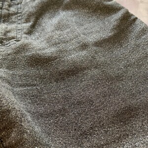 May include: Close-up of gray denim fabric with a subtle, shimmering effect. The fabric has a textured appearance, with visible stitching details near the pocket area. The overall tone is a muted gray with a hint of sparkle, suggesting a casual yet stylish garment.