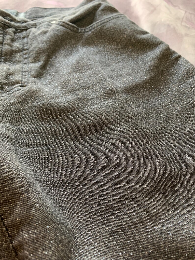 May include: Close-up of gray denim fabric with a subtle, shimmering effect. The fabric has a textured appearance, with visible stitching details near the pocket area. The overall tone is a muted gray with a hint of sparkle, suggesting a casual yet stylish garment.
