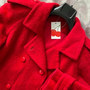 Peut inclure: Un manteau rouge vif à double boutonnage avec un col classique et des boutons. Le manteau est suspendu à un cintre noir, avec une étiquette représentant des cerises. Le tissu du manteau a un aspect doux et texturé.