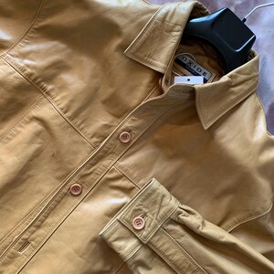 Puede incluir: Una camisa de cuero color canela con cuello y cierre de botones. La camisa tiene mangas largas con puños abotonados. La etiqueta de la marca dice "OXIDE SPORT". La camisa está colgada en una percha negra.