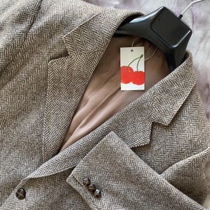 Peut inclure: Un blazer marron à chevrons de conception classique. Le blazer a un revers cranté, trois boutons sur le poignet et deux boutons sur le devant. Une étiquette avec une illustration de cerise est suspendue au col.
