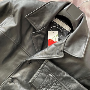 May include: A black leather jacket with a classic collar and button closure. The jacket is hanging on a black hanger. A tag with a cherry design is visible. The label reads "Good & Christensen Couture."