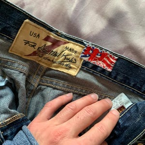 May include: Close-up of denim jeans with a leather tag that reads "USA For All Man Kind". The jeans have a faded wash and a small American flag tag. The jeans are size 24.