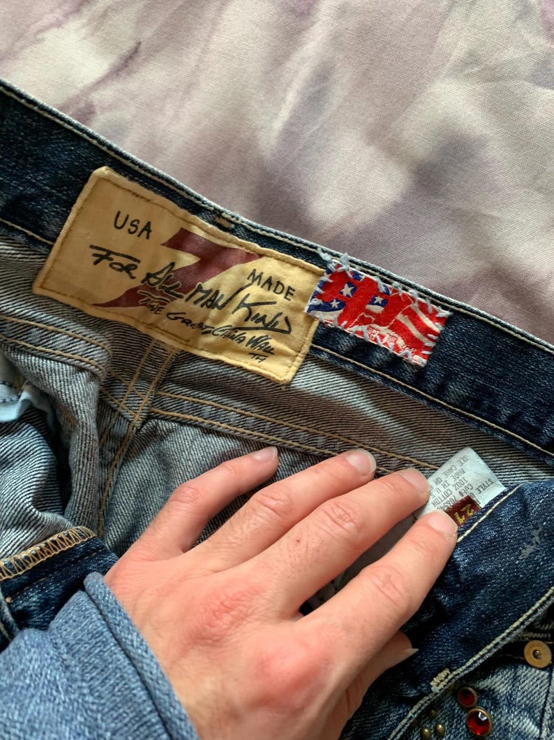 May include: Close-up of denim jeans with a leather tag that reads "USA For All Man Kind". The jeans have a faded wash and a small American flag tag. The jeans are size 24.
