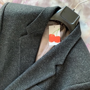 May include: A dark gray blazer with a textured fabric, hanging on a black hanger. The blazer has a notched lapel and a light brown lining. A tag with a red cherry design is attached to the hanger.