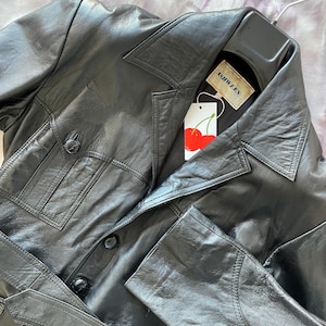 May include: Black leather jacket with a notched collar, two front pockets, and a belt. The jacket has a tag that says "ASHWEEN" and a cherry design.
