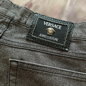 May include: Close-up of gray denim jeans with a black leather patch. The patch features the Versace logo and the words "JEANS COUTURE". The jeans have a classic five-pocket design and visible stitching.
