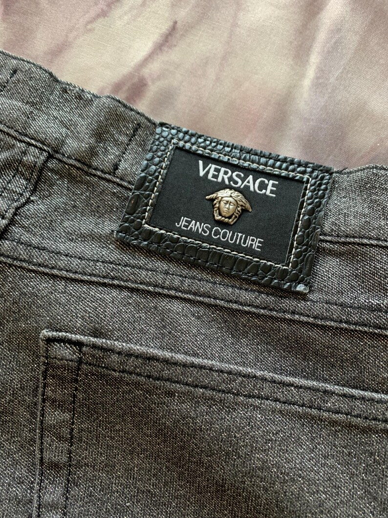 May include: Close-up of gray denim jeans with a black leather patch. The patch features the Versace logo and the words "JEANS COUTURE". The jeans have a classic five-pocket design and visible stitching.
