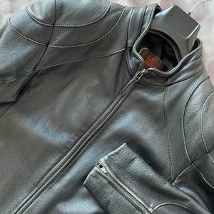 Peut inclure: Veste en cuir noir avec fermeture éclair et col montant. La veste présente des panneaux matelassés aux épaules et aux bras. La veste est suspendue à un cintre noir.