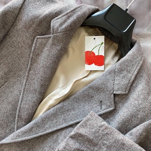 May include: A brown herringbone blazer with a notched lapel and a two-button closure. The blazer has a light-coloured lining and a tag with two red cherries. The blazer is hanging on a black hanger.